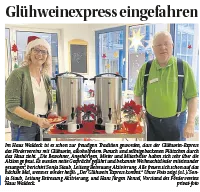 Zwei Personen in einem festlich geschmückten Innenraum stehen an einem Getränkewagen mit zwei großen Warmhaltebehältern; davor stehen Tassen und rote Weihnachtssterne, im Hintergrund sind Fenster mit Stern-Deko zu sehen.