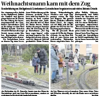 Mehrere Familien und Kinder stehen an einer Gartenbahnanlage im Freien und schauen den Modellzügen zu; im Vordergrund sind Gleise und kleine Landschaftsmodelle zu sehen, im Hintergrund Menschen in Winterkleidung vor Wohnhäusern.