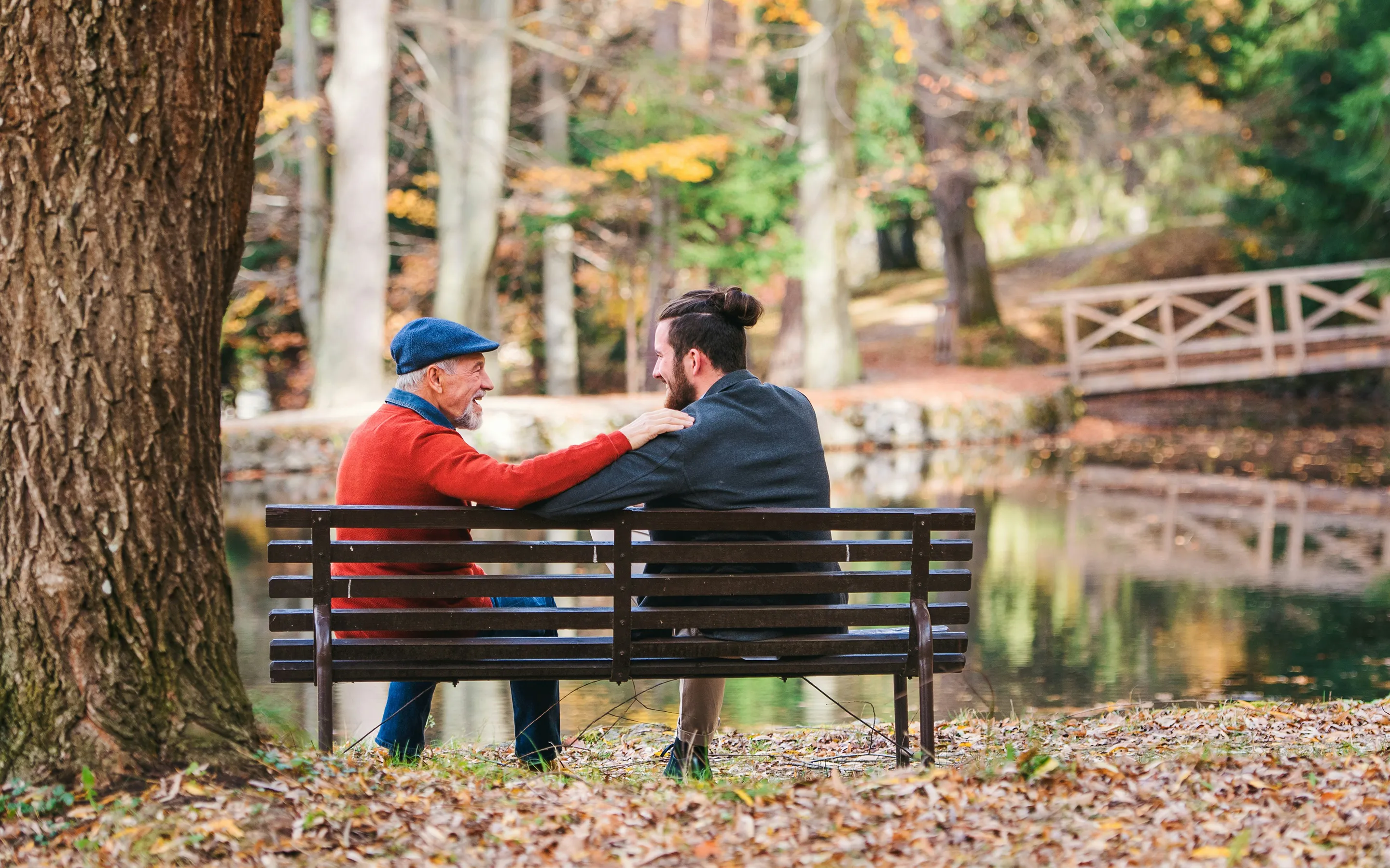 Zwei Männer auf einer Bank im herbstlichen Wald, ein jüngerer und ein älterer Mann, sie unterhalten sich freundschaftlich